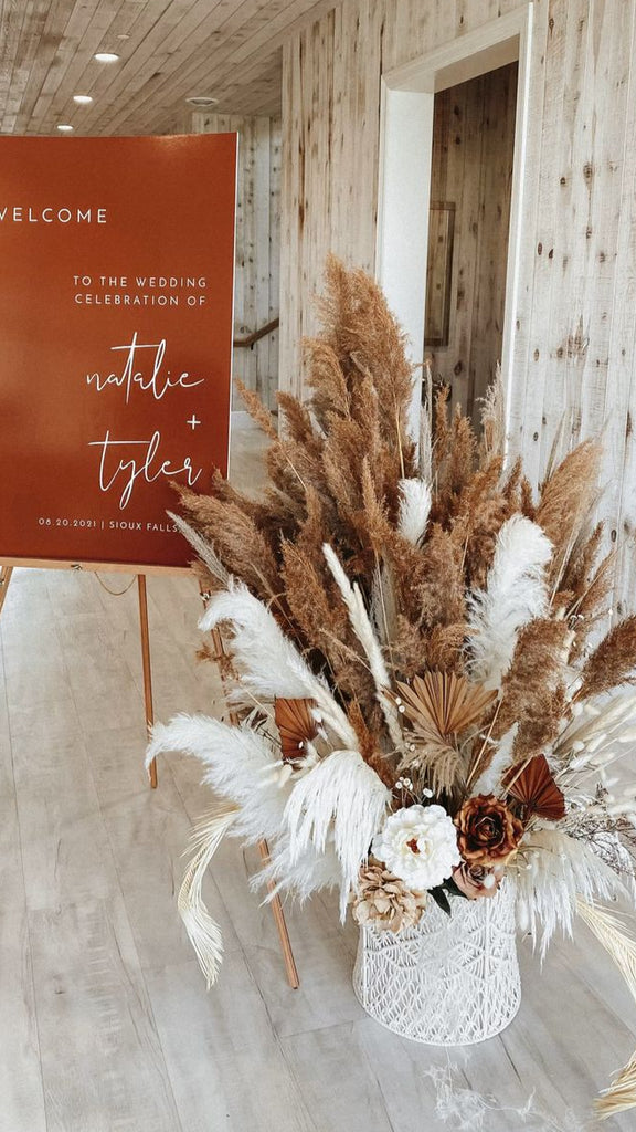 Decorative sign with 'Welcome to the wedding celebration of Natalie + Tyler' in a rustic setting with pampas grass and flowers.